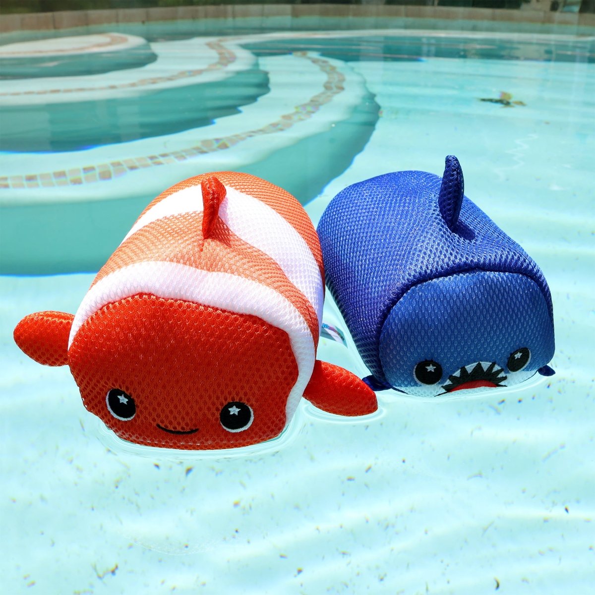Two Aqua Moosh-Moosh Crush plush pool toys float in clear water: one is orange and white like a smiling clownfish, the other is blue like a shark with a fin and cartoonish open mouth showing teeth.