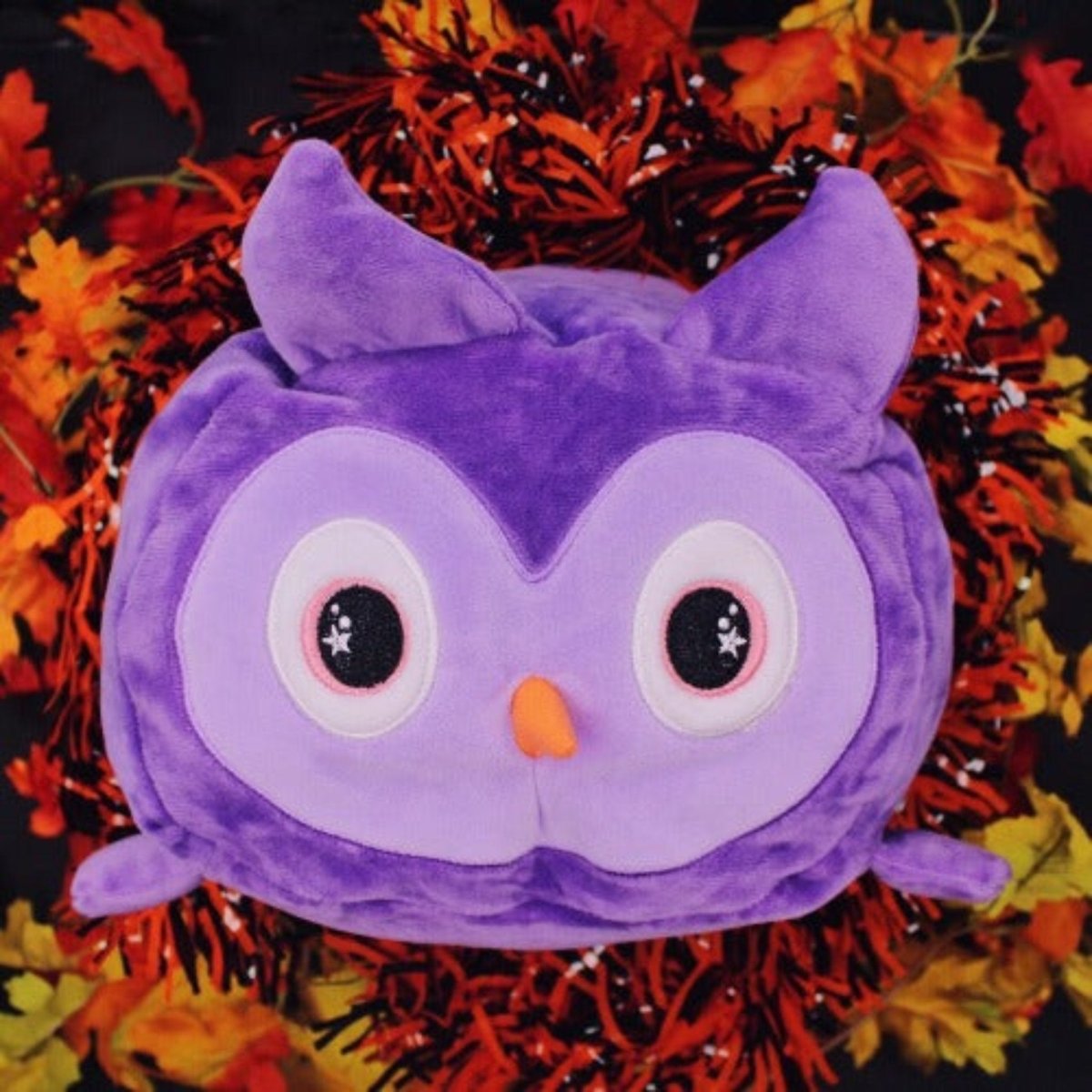 Archie the Owl Plushie, with big eyes and an orange beak, sits among faux autumn leaves and orange-black tinsel—making it the perfect Halloween plushie against a dark background.