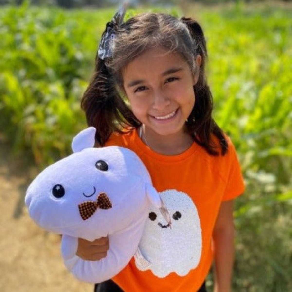 A young girl with dark hair tied with a bow smiles while hugging Boo the Ghost Plushie. She's wearing an orange shirt featuring a cartoon ghost. The background shows a sunny outdoor scene with greenery.