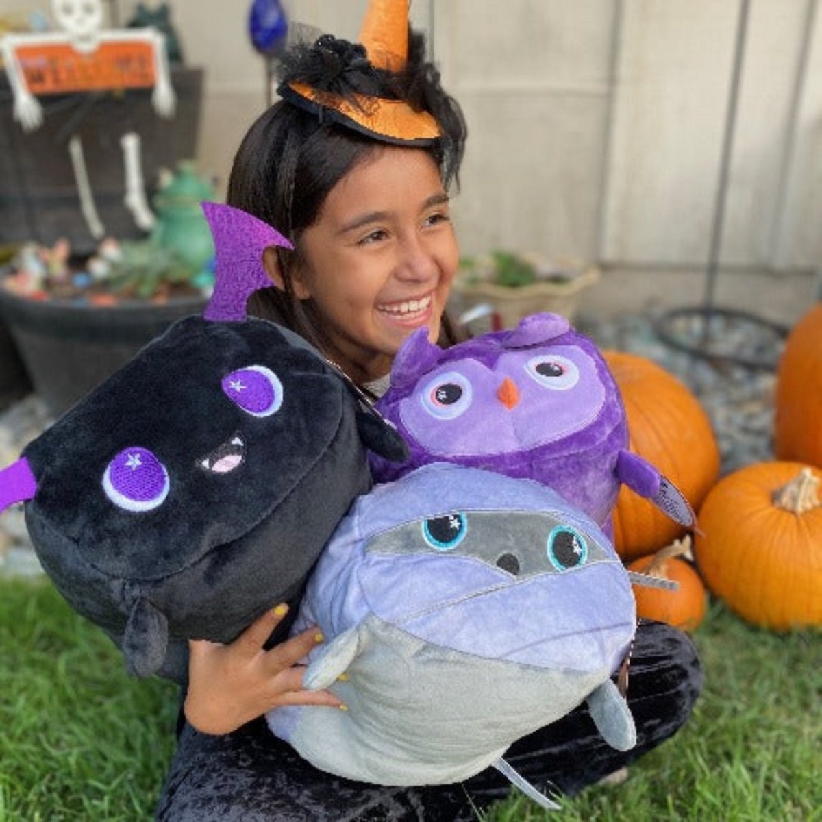 A child smiles holding three plushies: Cassie the Bat (black), a purple owl, and a gray mummy. Seated on grass with pumpkins, the child wears a witch hat headband amidst Halloween decorations.