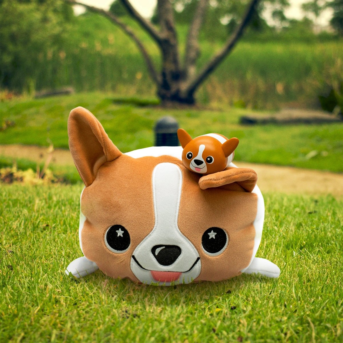 A large plush Corey the Puppy with floppy ears sits on the grass, while a small Corey the Puppy Vinyl Collectible Figurine perches playfully on its head. In the background, a tree and greenery appear softly blurred.