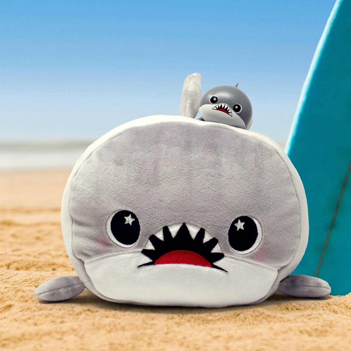 Crush the Shark, a cute vinyl collectible figurine with big eyes, sits by a surfboard on the sandy beach under a blue sky, making it perfect for ocean lovers.