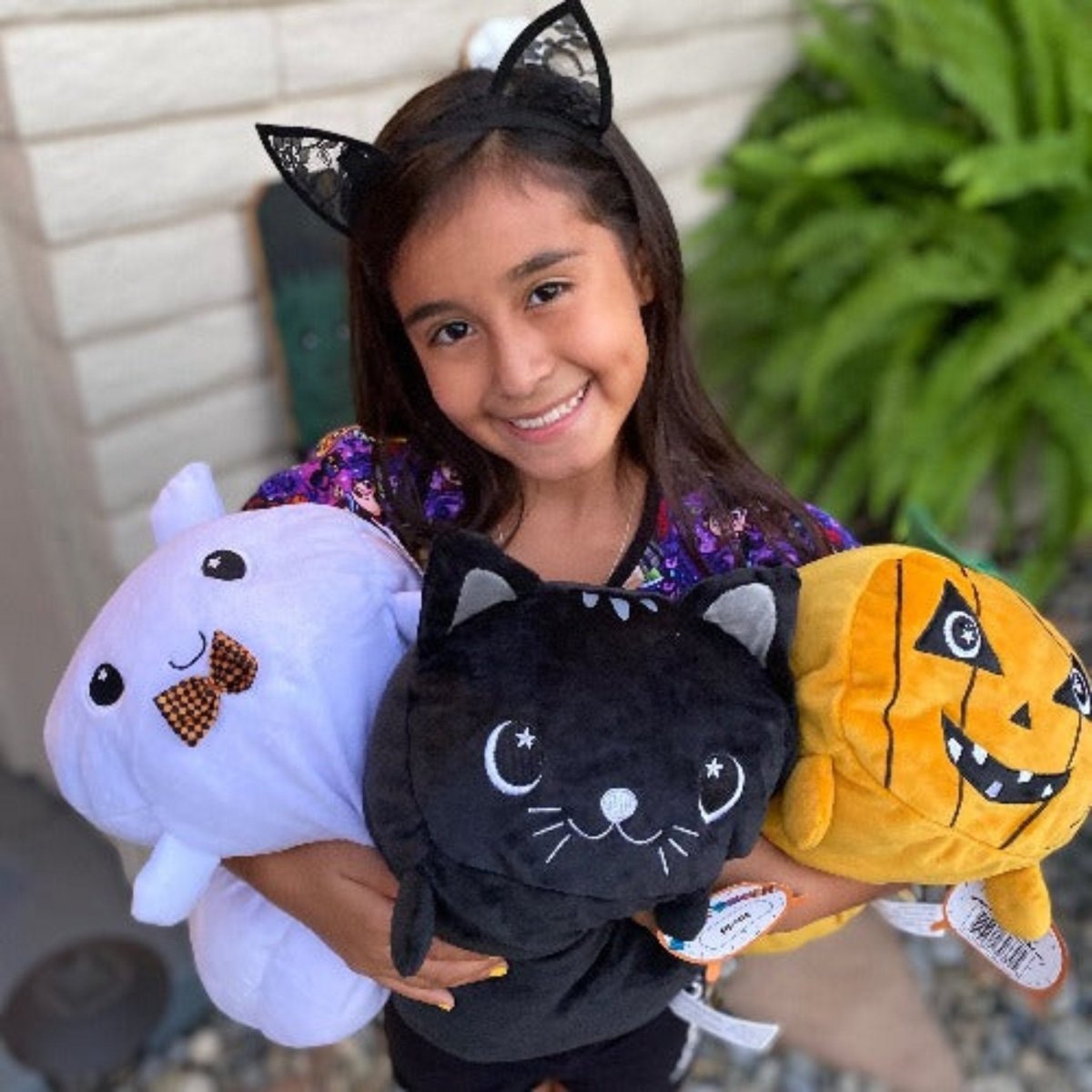 A young girl with cat ears smiles while holding three plushies: Fortune the Cat, a black cat, and an orange pumpkin with a face. Dressed in a sequined shirt, she stands outdoors.