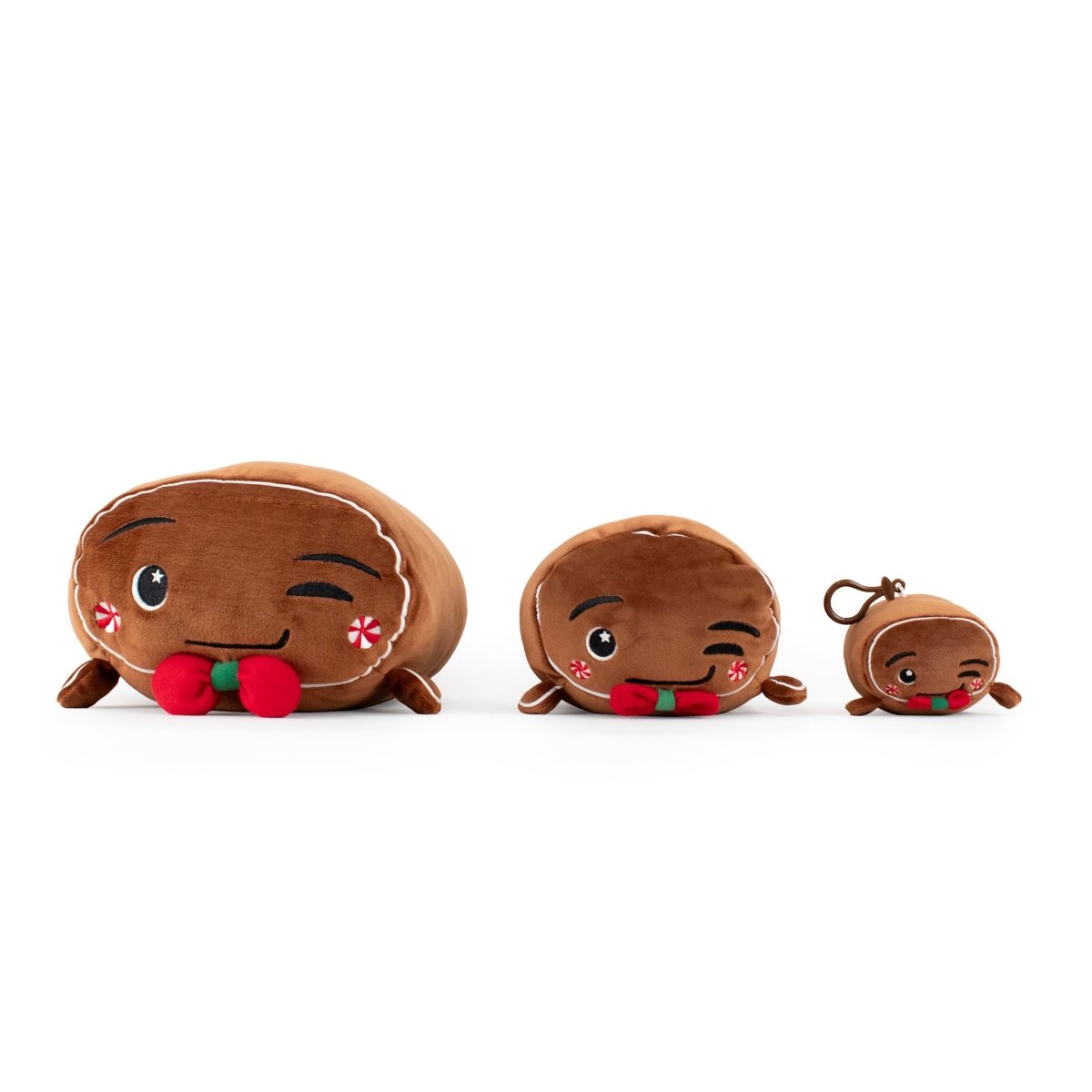 The Gingersnaps Clip-On features three plush gingerbread figures in varying sizes, each with a wink, rosy cheeks, festive bows, and white icing. The smallest is a keychain—ideal as a cute winter accessory or holiday plushie gift.