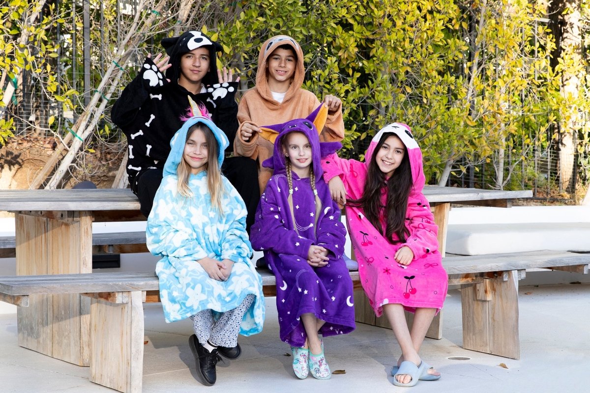 Five children sit on wooden benches outdoors, each smiling and wrapped in a colorful kids fleece blanket or the Hooded Blanket - Vee the Bee. They are surrounded by greenery, creating a relaxed and cheerful scene.