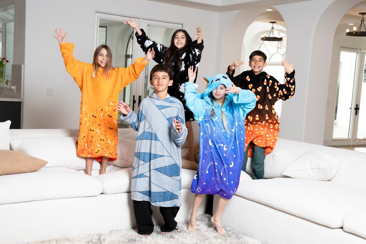 Five kids, each wearing a Vincent the Pumpkin Hooded Blanket, pose excitedly on a white sectional couch in a bright, modern living room.