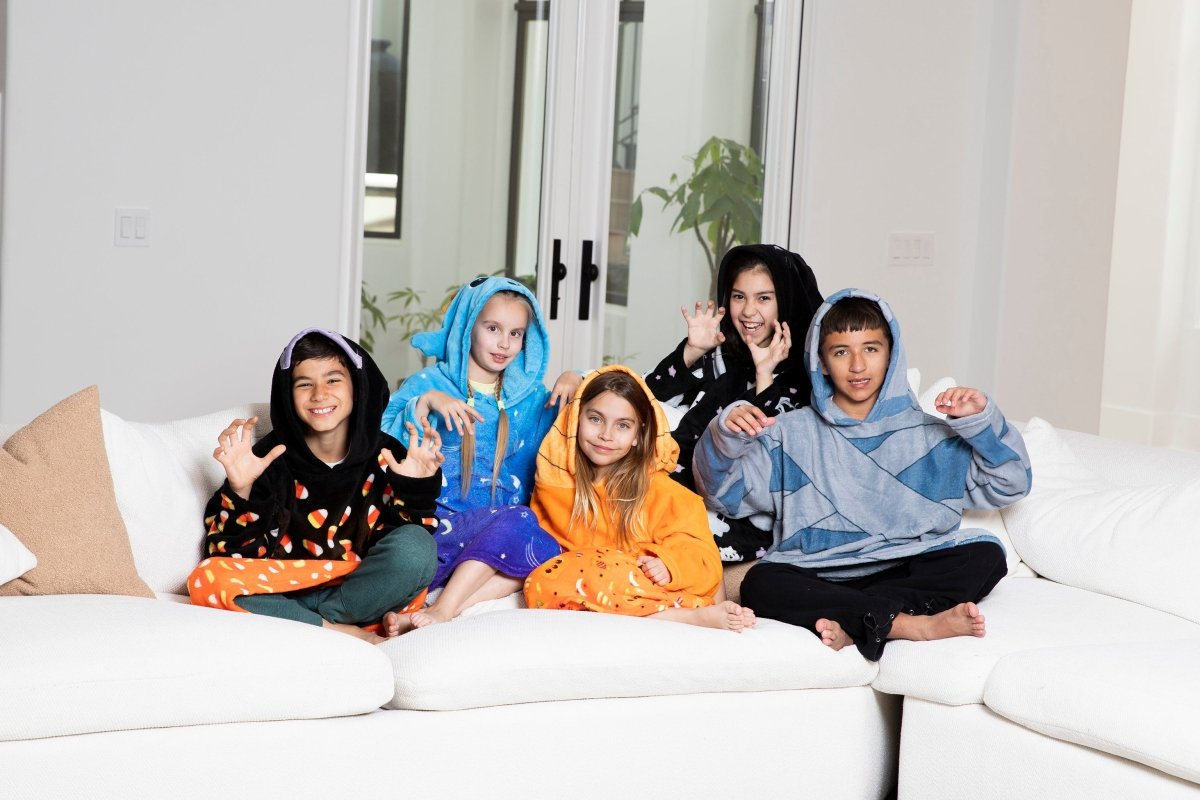 Five children smile and make playful claw gestures on a white couch, each wrapped in a Vincent the Pumpkin Hooded Blanket. The cozy, colorful fleece blankets stand out against the bright, modern room in the background.