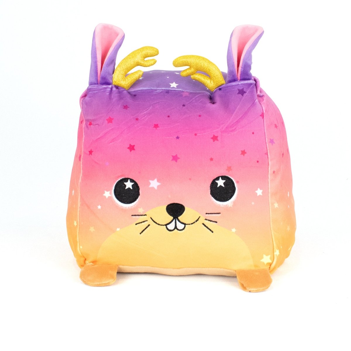 Jinx the Jackalope is a square, machine washable plush toy with a gradient pink, purple, and yellow body, star patterns, smiling face, rabbit ears, golden antlers, and small feet—perfect for plush fans. Pictured on a white background.