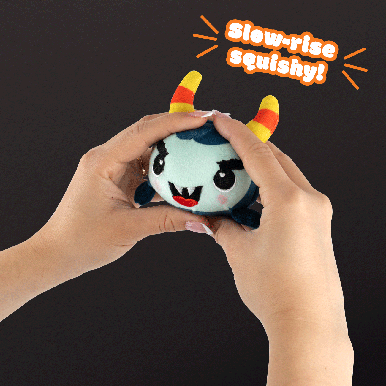 Two hands squish the Gretel Spooky Plushy Skwishy, a round blue monster plush with yellow and red striped horns and an angry face, against a black background. Text reads “Slow-rise squishy!” in orange and white.
