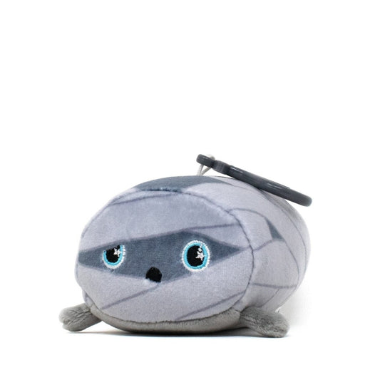 The Nile the Mummy Clip-On is a small, soft plush toy with big blue eyes, wrapped in light gray fabric. It's round and features a keychain attached to the top.