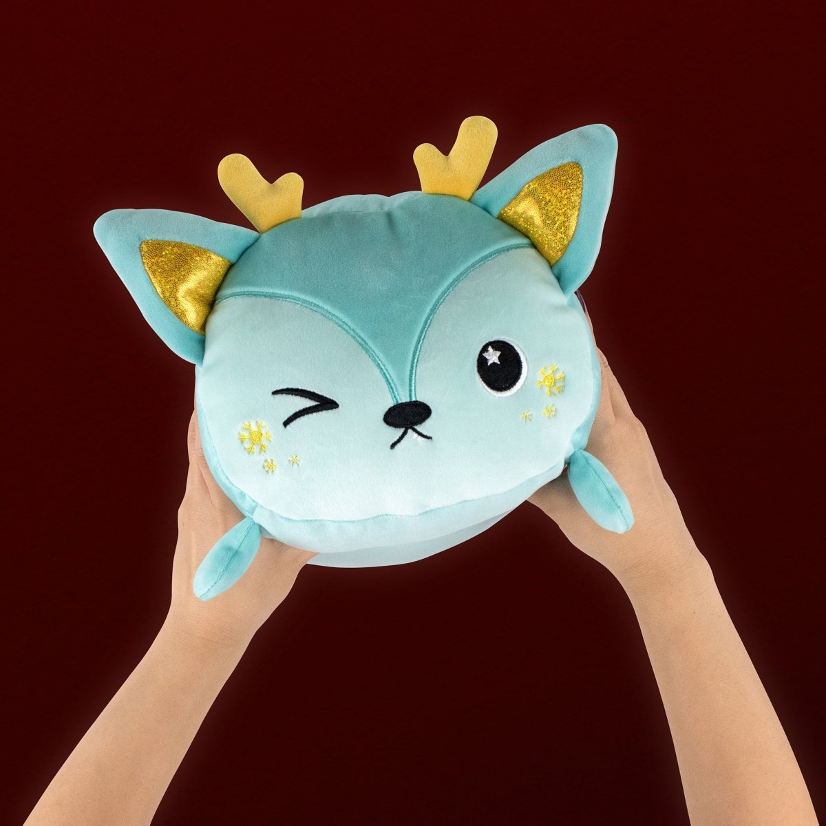 Two hands hold Blitzy the Deer, a round teal plushie with yellow antlers, golden inner ears, a winking face, and a star in one eye. This soft, cuddly stuffed animal pops against the dark red background—perfect for the holidays.