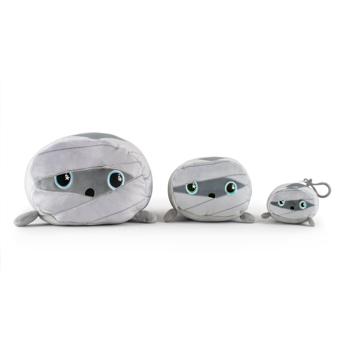 Nile the Mummy Plushies, arranged from largest to smallest, feature soft gray fabric and large round eyes for a friendly look. The smallest has a keychain.