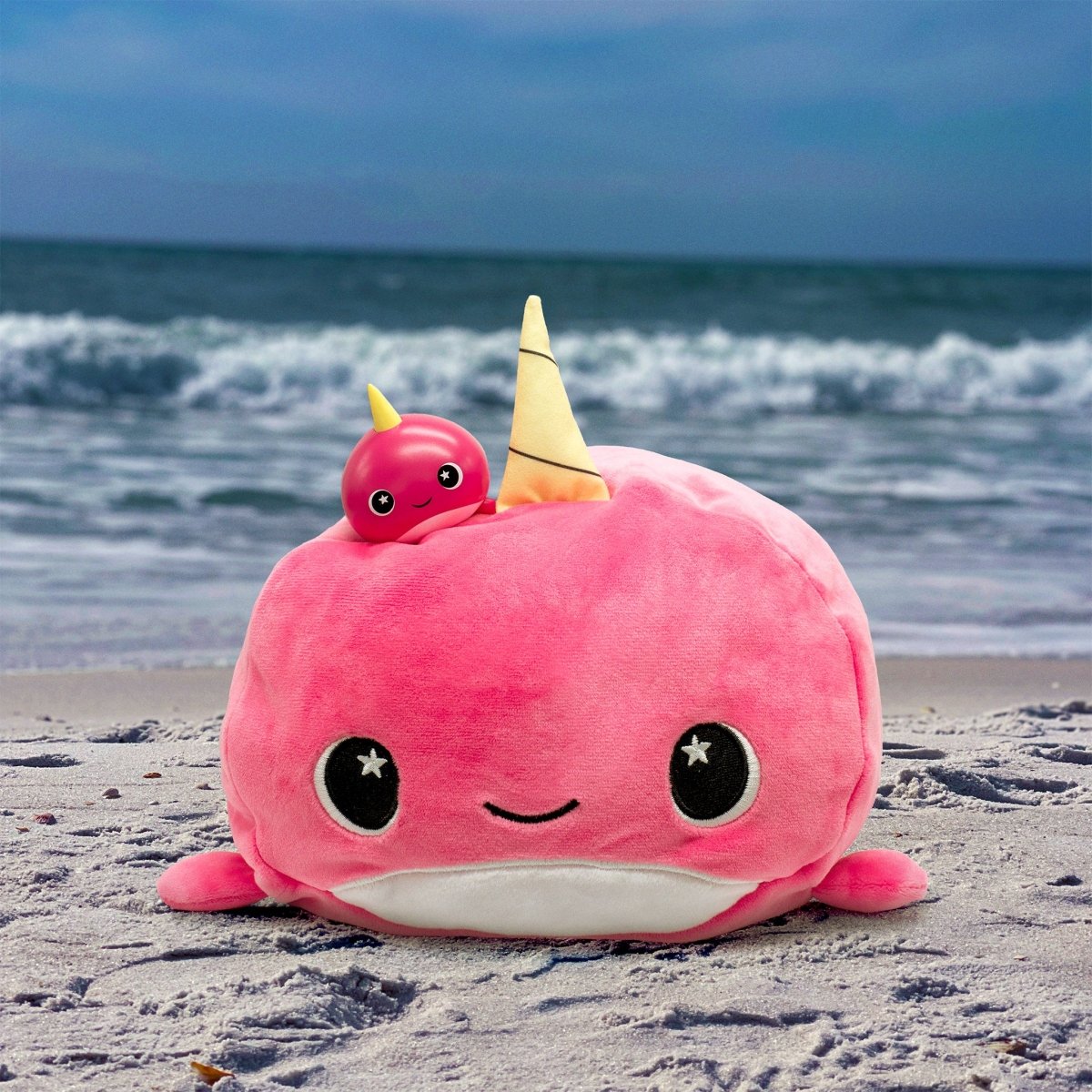 Bubblegum the Narwhal Vinyl Collectible Figurine, featuring a yellow horn and a smiling face, sits on a sandy beach near the ocean with waves and clouds, creating an adorable seaside scene. A smaller pink Moosh-Moosh figurine rests on top.