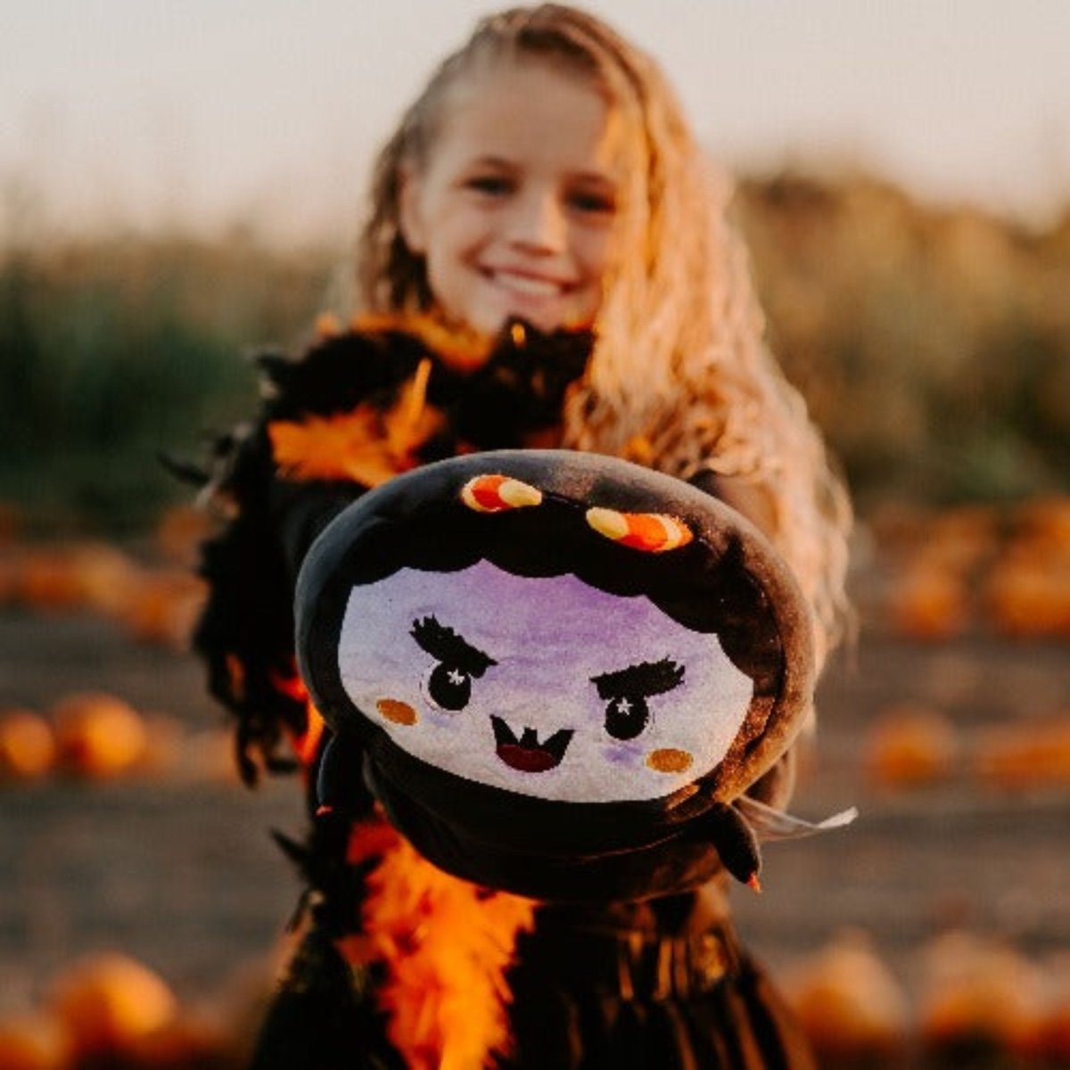 A smiling child with long blonde hair holds out a Fang the Monster Plushie with a cartoon vampire face while standing outdoors among scattered pumpkins.