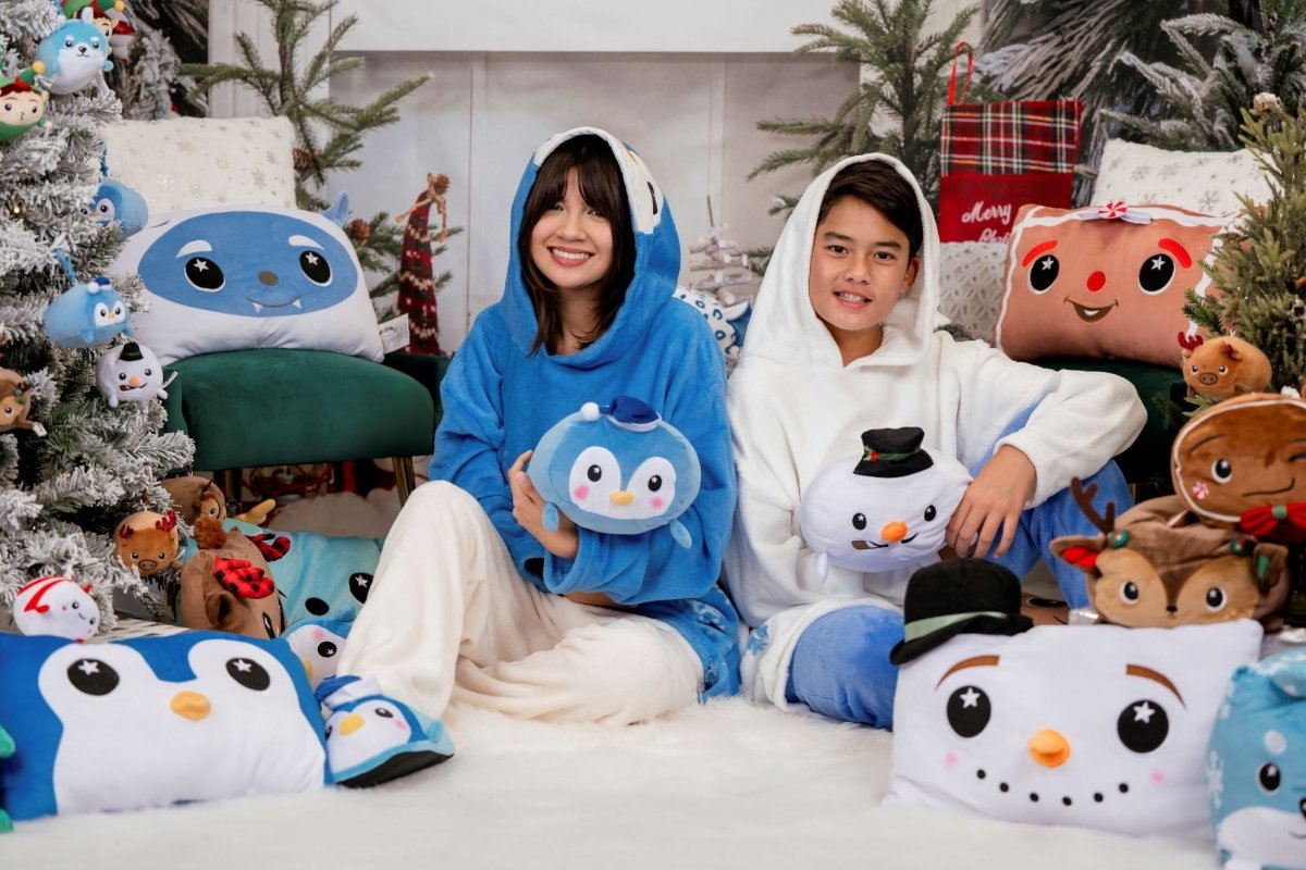 Two people in cozy outfits sit on a white rug surrounded by cute plush pillows and holiday-themed stuffed toys, like penguins and Jack the Snowman Hooded Blanket. Decorated Christmas trees and festive ornaments enhance the cheerful, festive atmosphere.