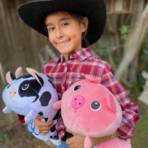 A child in a black cowboy hat and red plaid shirt smiles while holding ultra-soft plushies—a cow and Pinky the Pig Plushie—in their arms, standing outdoors with a wooden fence and greenery in the background.