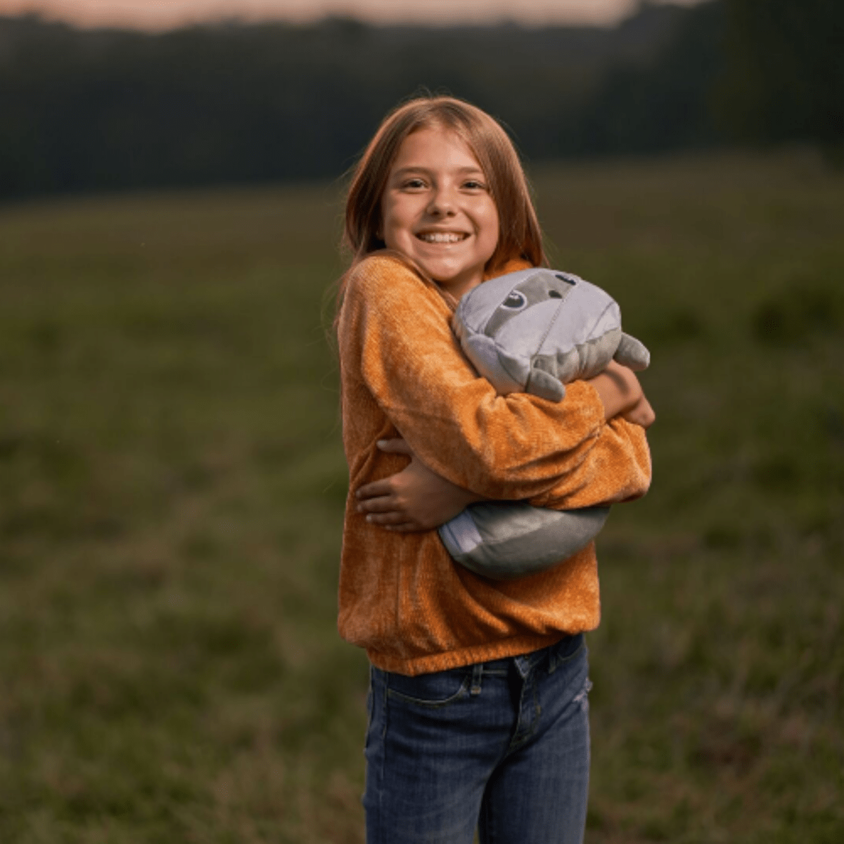 In a grassy field at dusk, a young girl smiles brightly, hugging Nile the Mummy Plushie. She's dressed in an orange sweater and blue jeans, with a blurred landscape of trees behind her.
