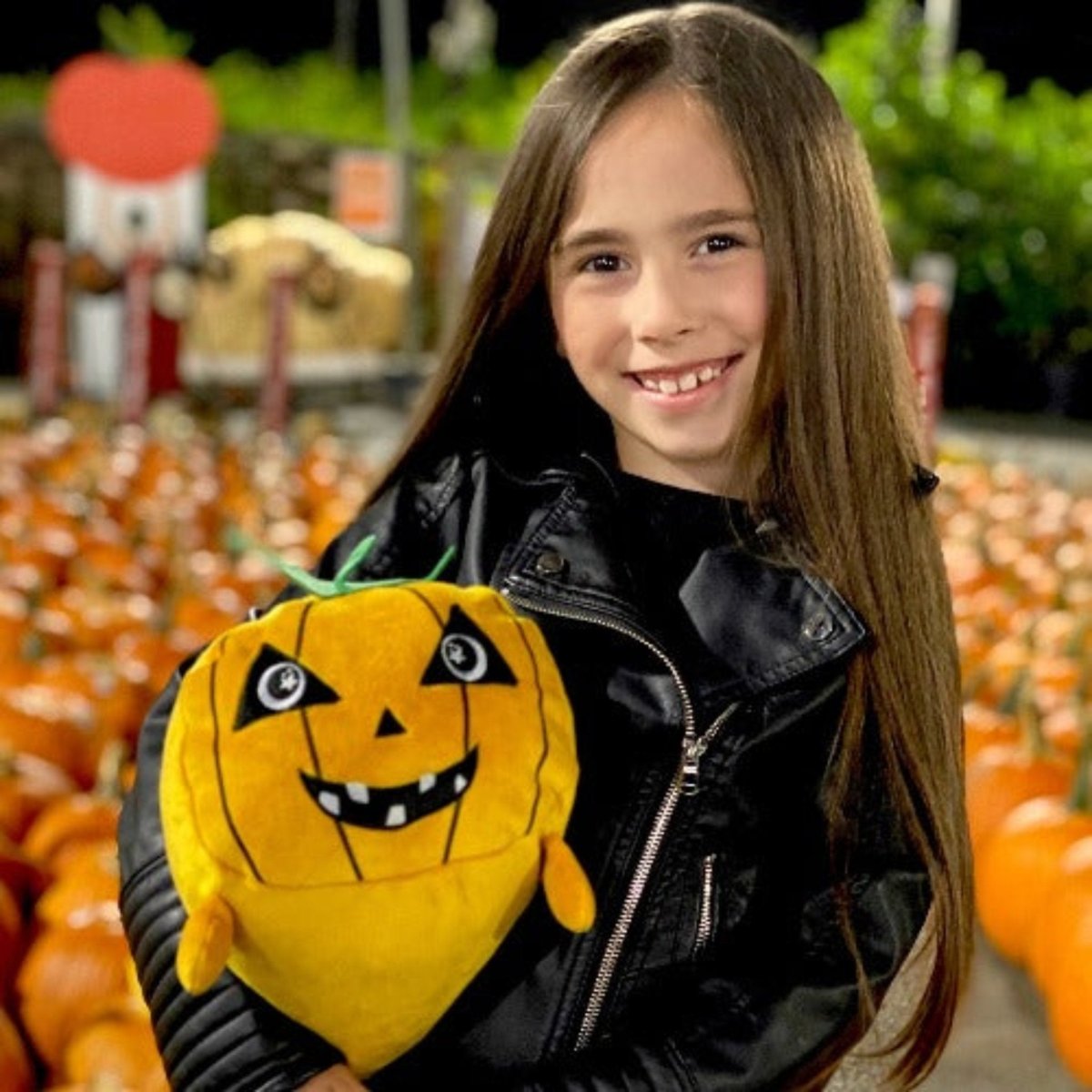 A smiling child with long brown hair in a black leather jacket holds Vincent the Pumpkin Plushie, a cuddly stuffed pumpkin with a happy face, surrounded by orange pumpkins in a pumpkin patch.