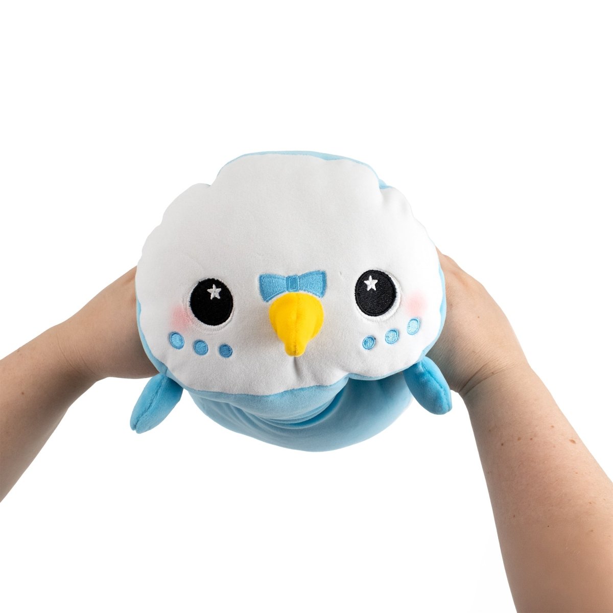 Two hands hold Kiki the Parakeet, a plush toy made from ultra-soft fabric, showing its blue and white body, big eyes, yellow beak, and small wings against a plain white background.