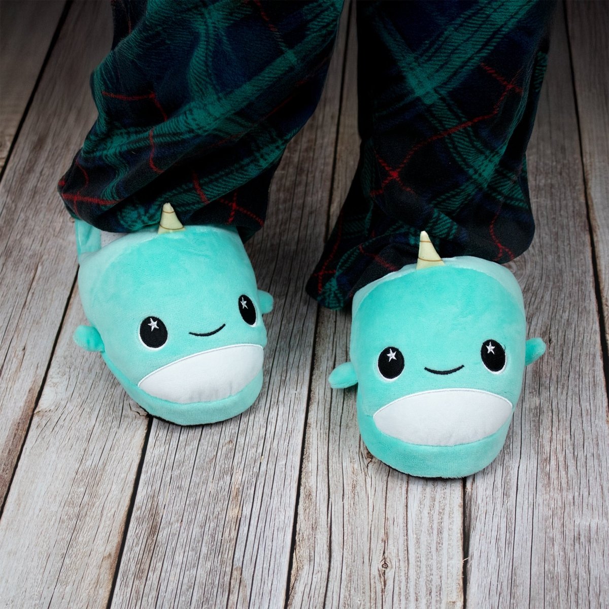 A child wears Chunky Monkey Kids Slipper with smiling faces, little horns, star-shaped eyes, and non-slip soles alongside dark plaid pajama pants on a wooden floor—perfect for cozy comfort at home.