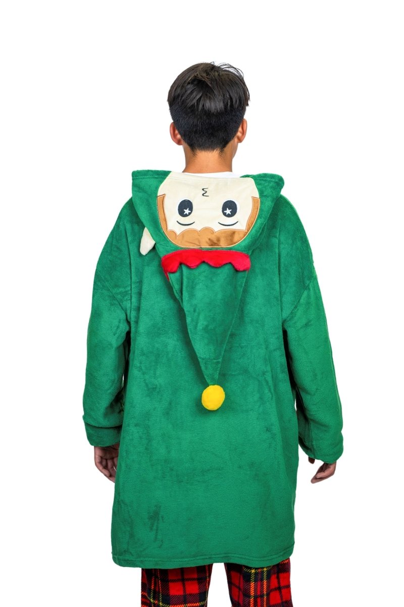 A person is seen from behind wearing the Jingle the Elf Hooded Blanket, featuring a green design with a Christmas tree and cartoon face on the hood. The outfit includes a soft yellow pom-pom, red accents, and is paired with cozy red plaid pants.