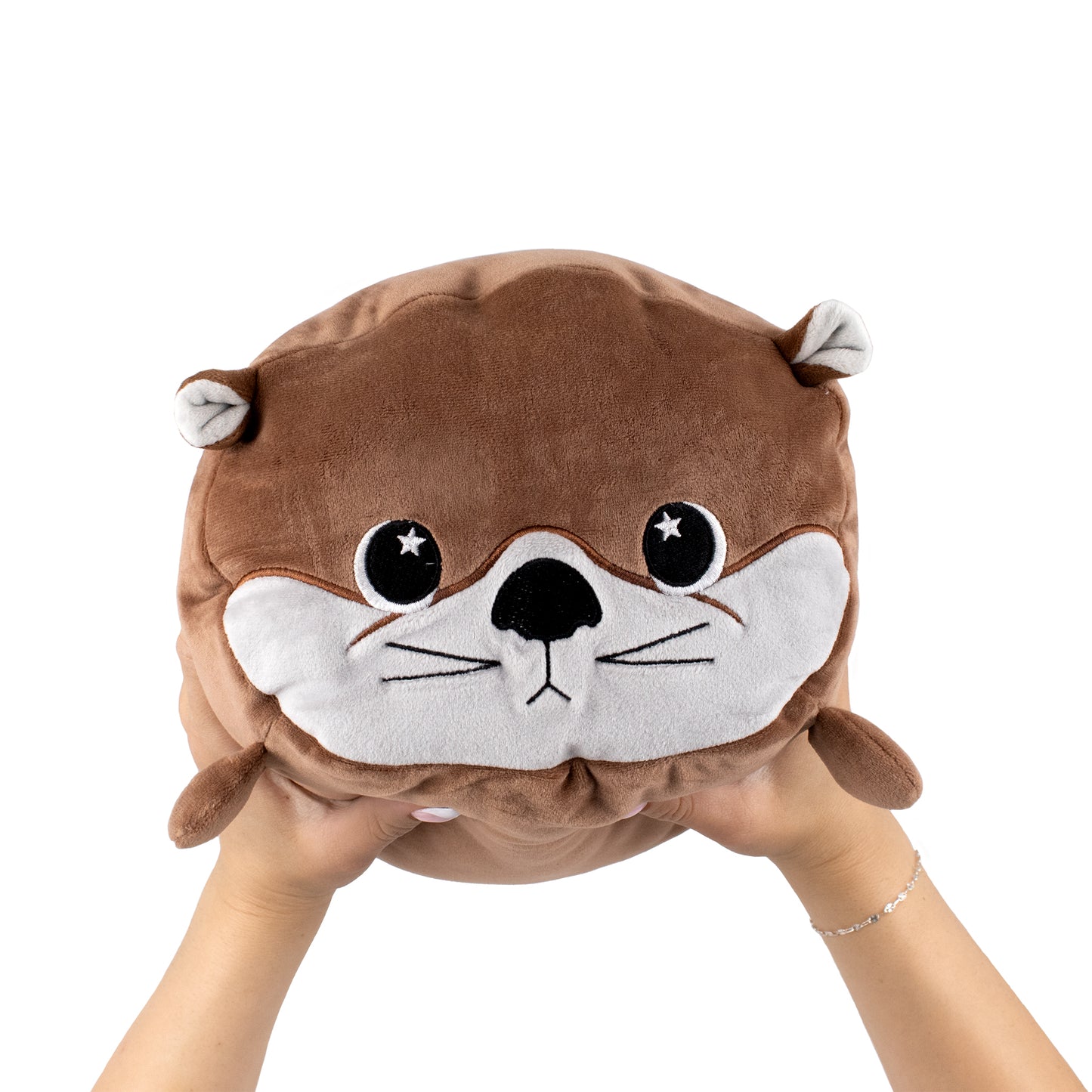 Two hands hold up Slick the Otter, a round brown plush toy with a cute face, small ears, big eyes, black nose, and embroidered whiskers, against a plain white background.