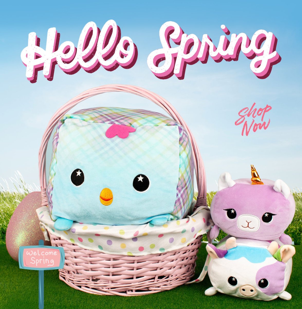A pastel pink basket holds a cute cube plush with a smiling face, surrounded by two animal plushies. Text reads "Hello Spring" and "Shop Now" against a grass and sky background with a decorated egg.