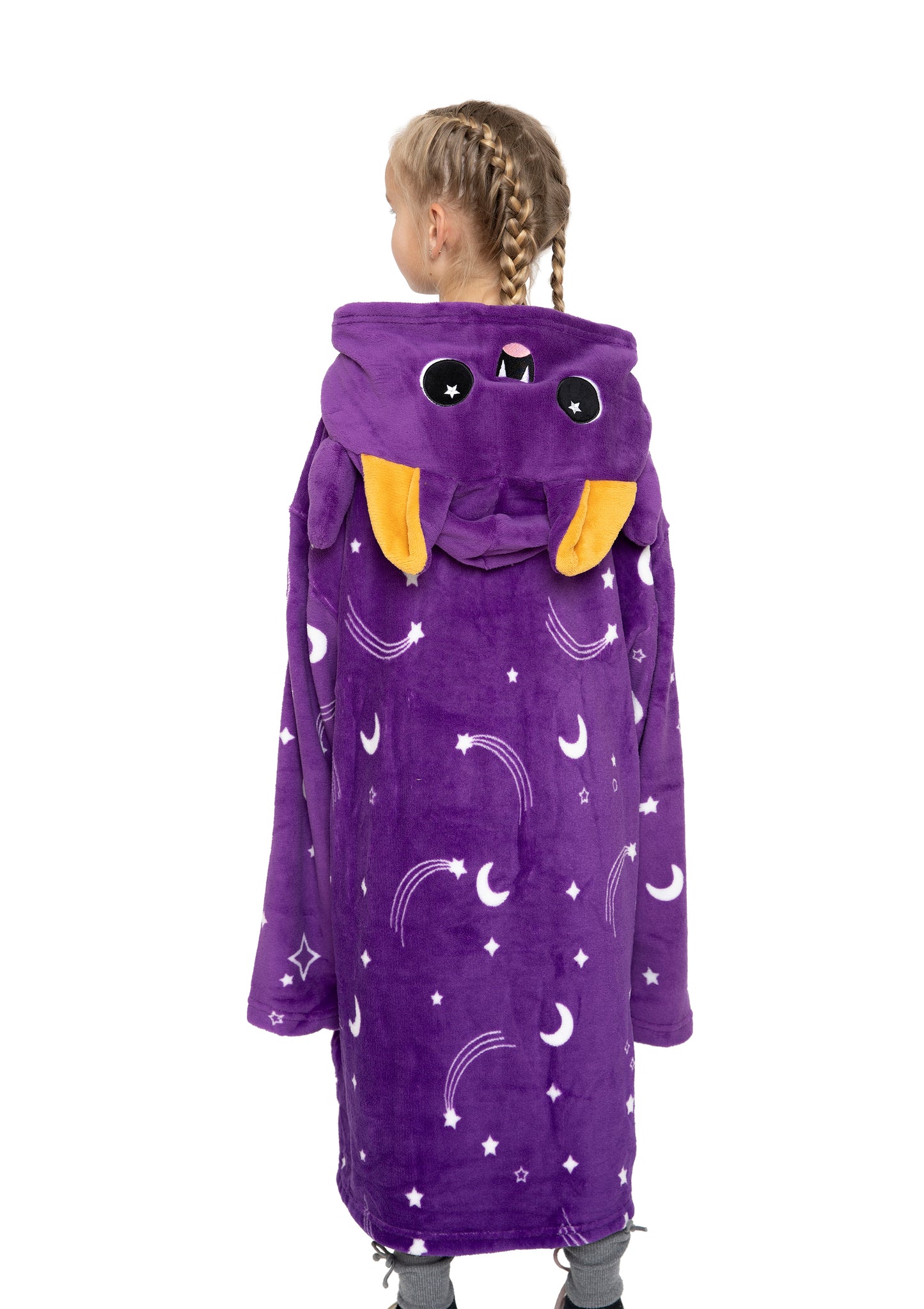A child with braided hair wears the Oswald the Bat Hooded Blanket—a long, plush purple hoodie decorated with moons, stars, and rainbows. The hood features a cute bat face with big eyes and orange ears. The child faces away from the camera.