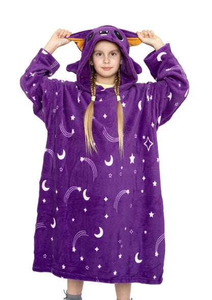 A young girl wears the Hooded Blanket - Oswald the Bat, featuring a purple design with moon and star patterns, cat-like ears, and a cute face. She faces forward, showing her long blond hair styled in braids.