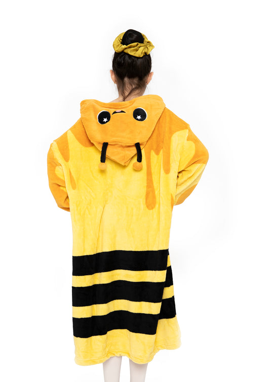 A person with a yellow hair scrunchie faces away, wearing the Hooded Blanket - Vee the Bee: a fuzzy yellow robe with black stripes and a hood featuring a cute cartoon bee face. The background is plain white.