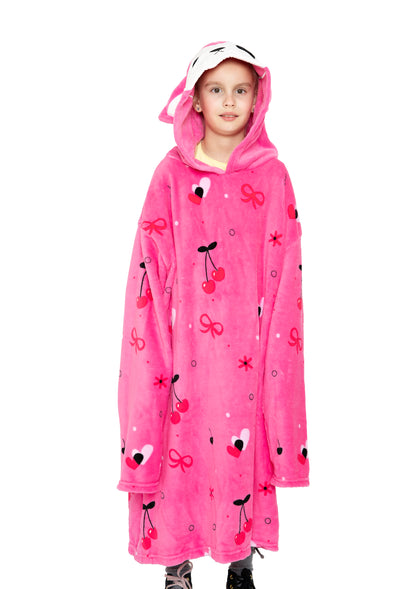 A young child stands against a white background wearing the oversized, bright pink "Hooded Blanket - Doe the Deer," featuring cherry and bow patterns and a white animal face on the hood.
