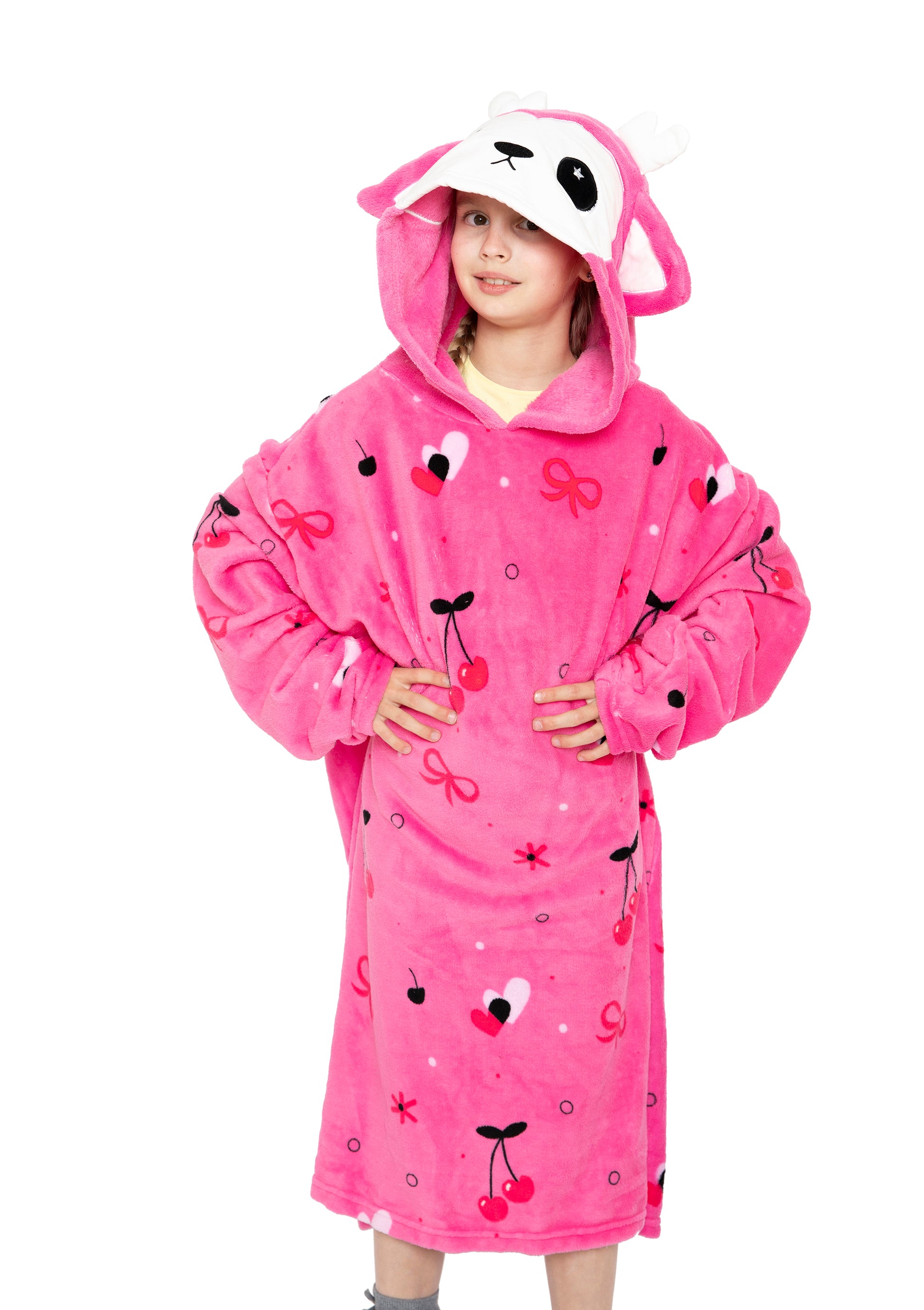 A young girl, hands on hips and smiling, wears the Hooded Blanket - Doe the Deer in bright pink with antlers, ears, and an animal face. The kids’ fleece blanket features heart, bow, and cherry designs.