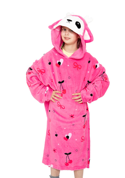 A young girl, hands on hips and smiling, wears the Hooded Blanket - Doe the Deer in bright pink with antlers, ears, and an animal face. The kids’ fleece blanket features heart, bow, and cherry designs.