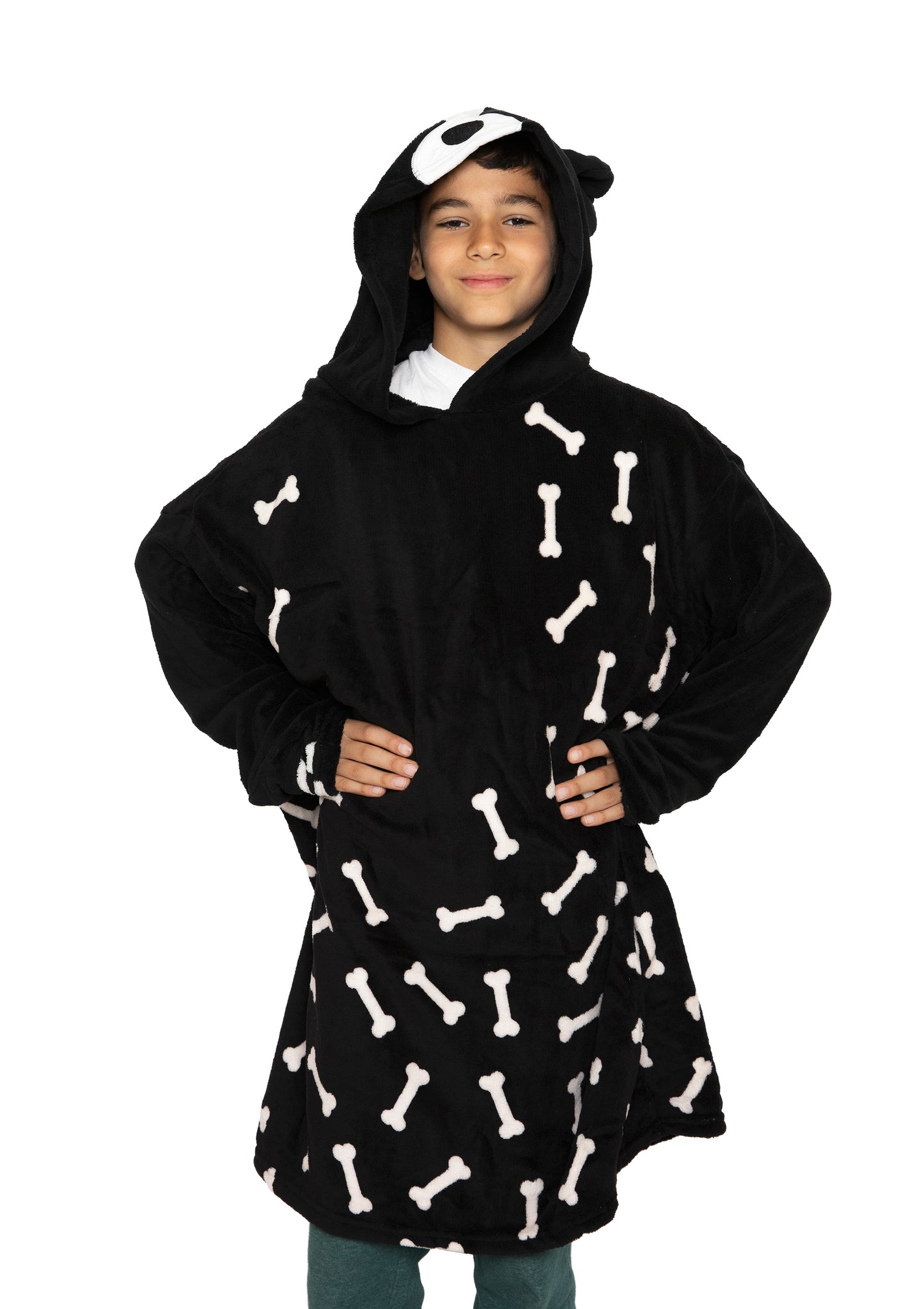 A smiling child stands hands-on-hips against a white background, wearing the Hooded Blanket - Ollie the Corgi, a black kids' hooded blanket featuring white bone patterns and a playful dog face on the hood.
