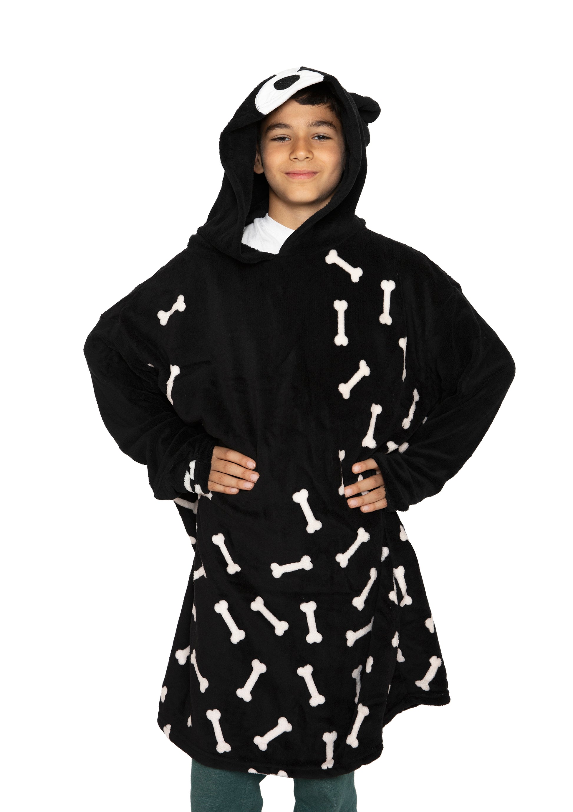 A smiling child stands hands-on-hips against a white background, wearing the Hooded Blanket - Ollie the Corgi, a black kids' hooded blanket featuring white bone patterns and a playful dog face on the hood.
