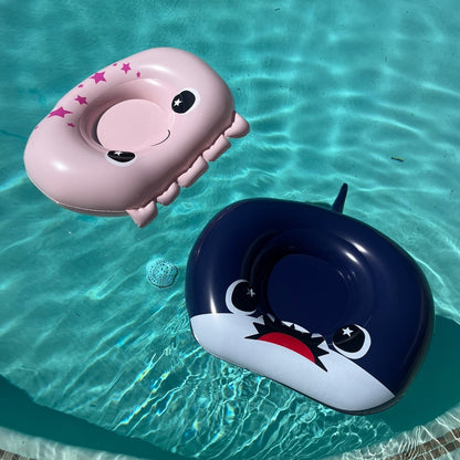 A pink Moosh-Moosh pool float with a smiling face and stars, and a navy blue Neptune 32" Inflatable Pool Float shaped like a shark, drift together in a clear blue swimming pool.