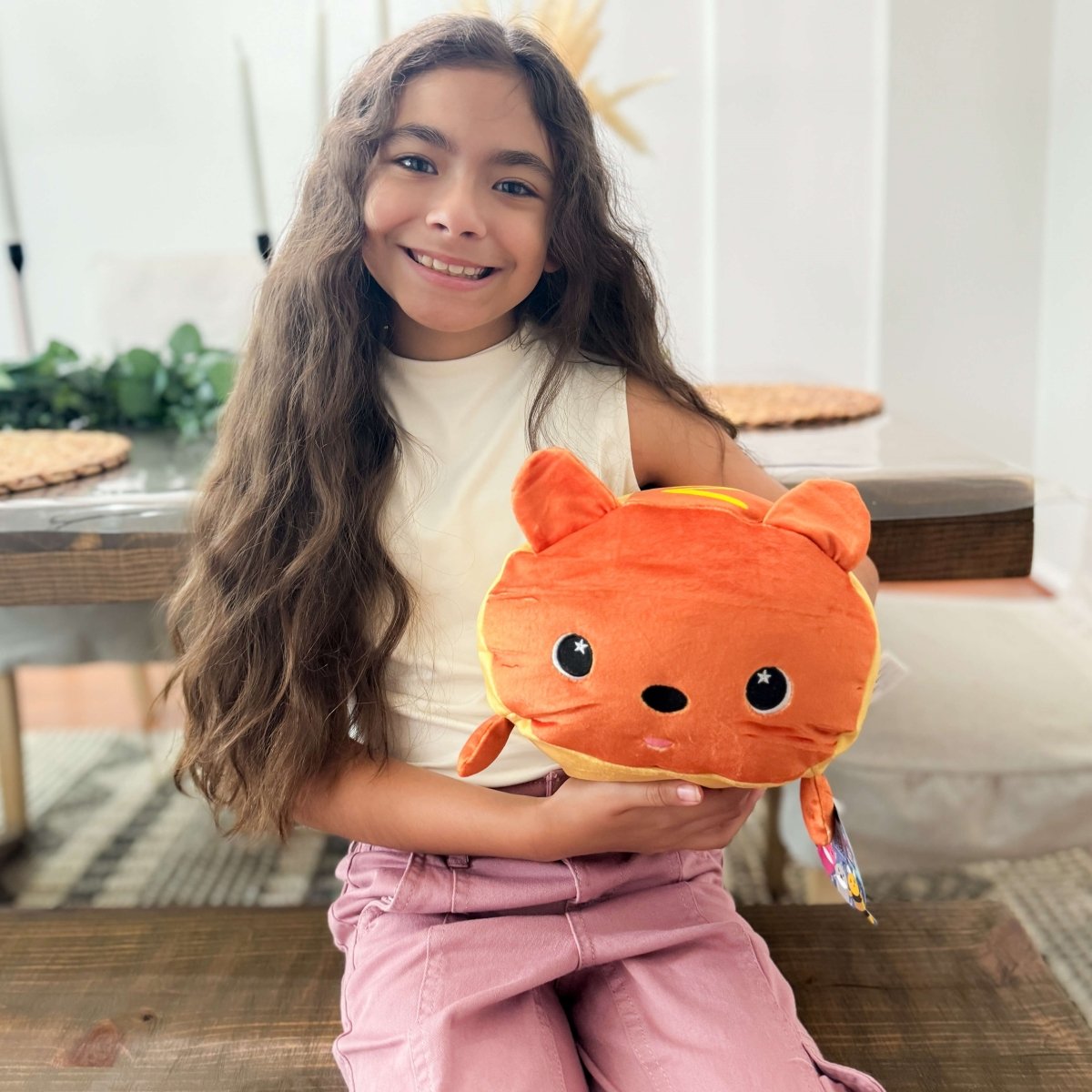A young girl with long brown hair smiles while sitting at a table, cuddling Gilbert the Hotdog Plushie in her arms. The room is brightly lit, and lush greenery is visible in the background.