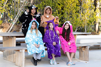 Five children sit on wooden benches outdoors, each smiling and wrapped in a colorful kids fleece blanket or the Hooded Blanket - Vee the Bee. They are surrounded by greenery, creating a relaxed and cheerful scene.