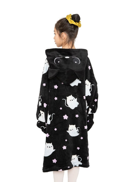 A girl with a yellow scrunchie wears the Fortune the Cat Hooded Blanket, a black fleece robe with white cartoon cats and purple stars. The hood has cat ears and a face. She is turned away, displaying the cozy blanket’s back.