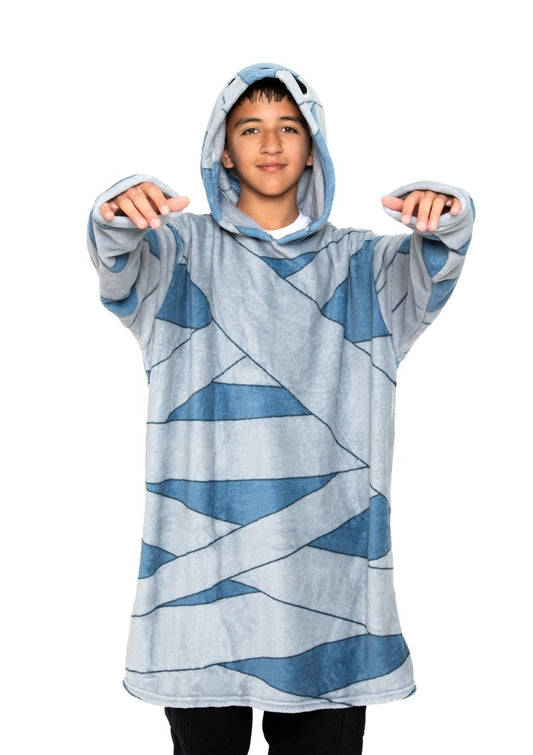 A boy wears the Nile the Mummy Hooded Blanket—an oversized blue and gray fleece with a mummy wrap pattern—and stands with arms raised and bent forward like a mummy against a white background.