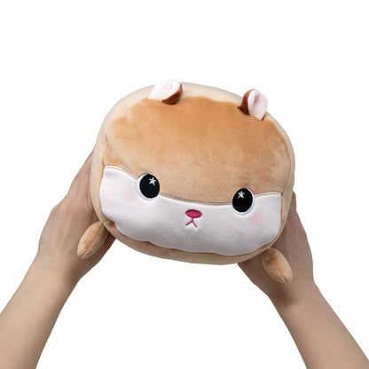 A pair of hands holds Hammy the Hamster, a round brown and white plush toy with large black eyes, small ears, and a cute pink nose, against a plain white background.