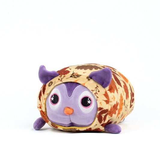 The Archie Spooky Slumber Plushie features a round, purple face with big eyes and small ears, crafted from ultra-soft materials and wrapped in a tan blanket adorned with brown and orange autumn leaves.