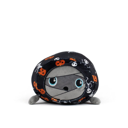 The Nile Spooky Slumber Plushie is a round, ultra-soft toy mummy with big blue eyes, wrapped in a black blanket featuring orange pumpkins, white skulls, and bones. This adorable plush sits on a white background.