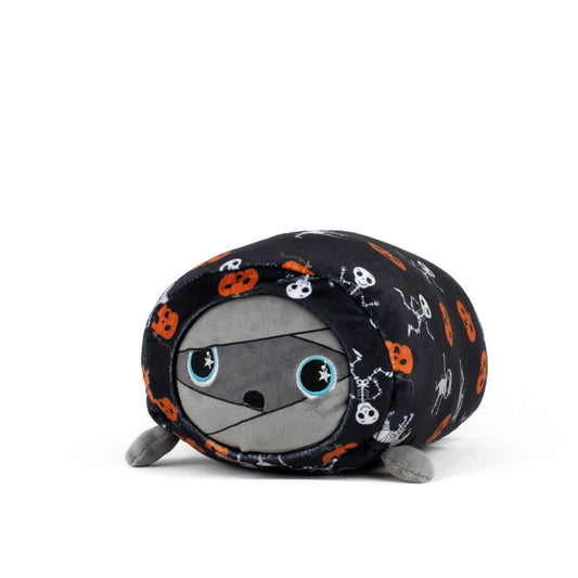 The Nile Spooky Slumber Plushie is a mummy-shaped toy made from ultra-soft materials, wrapped in black fabric with orange pumpkins and white skeletons. It has large blue eyes, a gray face, and a cute, round look.