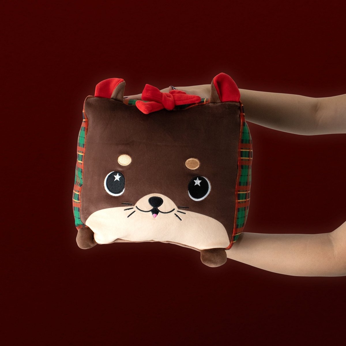 A pair of hands holds Blitz the Polar Bear, a square, ultra-soft plushie with a cute brown animal design, red bow, plaid sides, round eyes, and a cream-colored face, set against a dark red background.