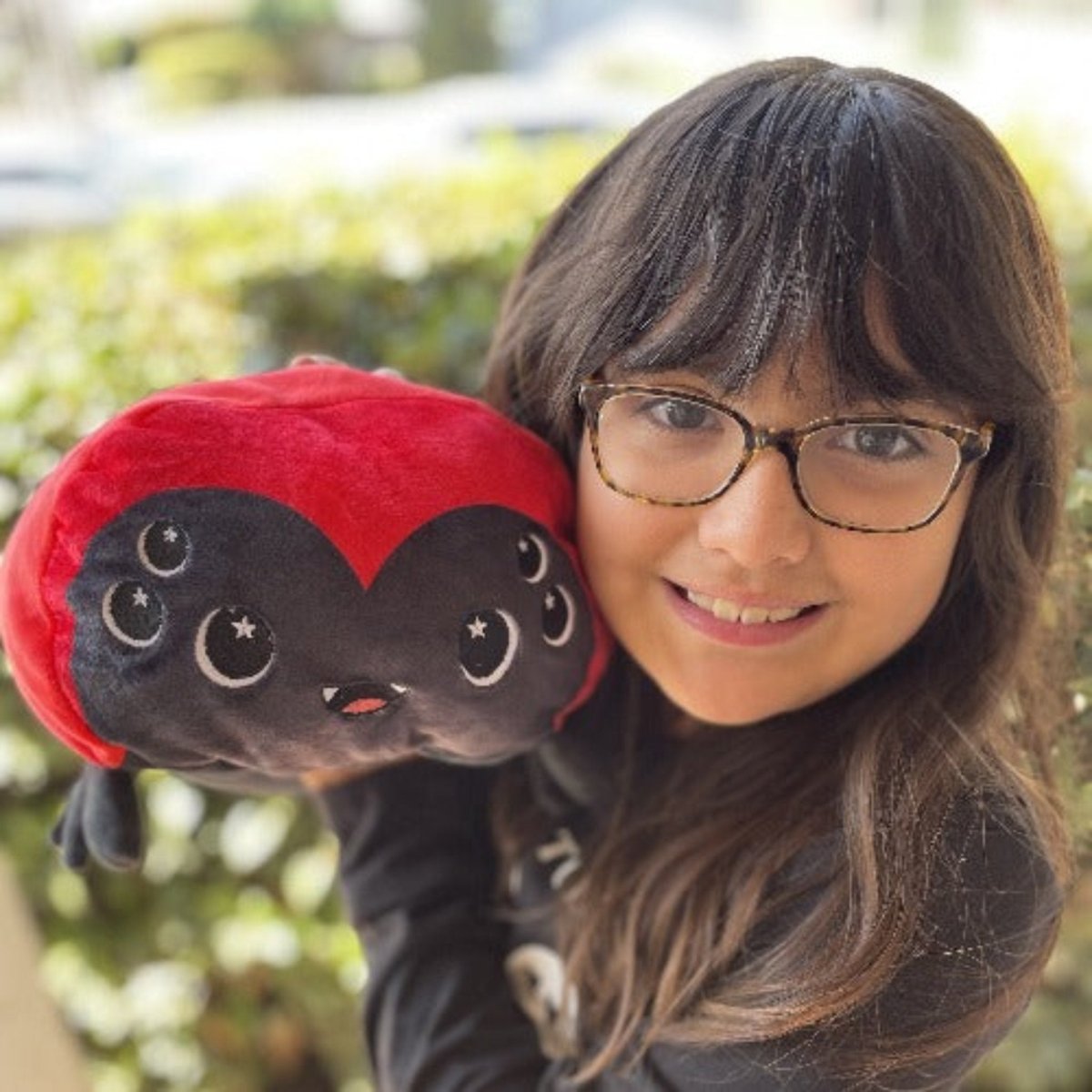 A smiling child with glasses holds a Muffet the Spider Plushie—a cute black and red heart-shaped toy with starry eyes—while green foliage is blurred in the background.