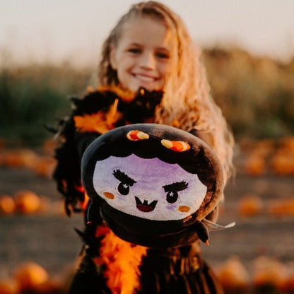 A smiling child with long blonde hair holds out a Fang the Monster Plushie with a cartoon vampire face while standing outdoors among scattered pumpkins.