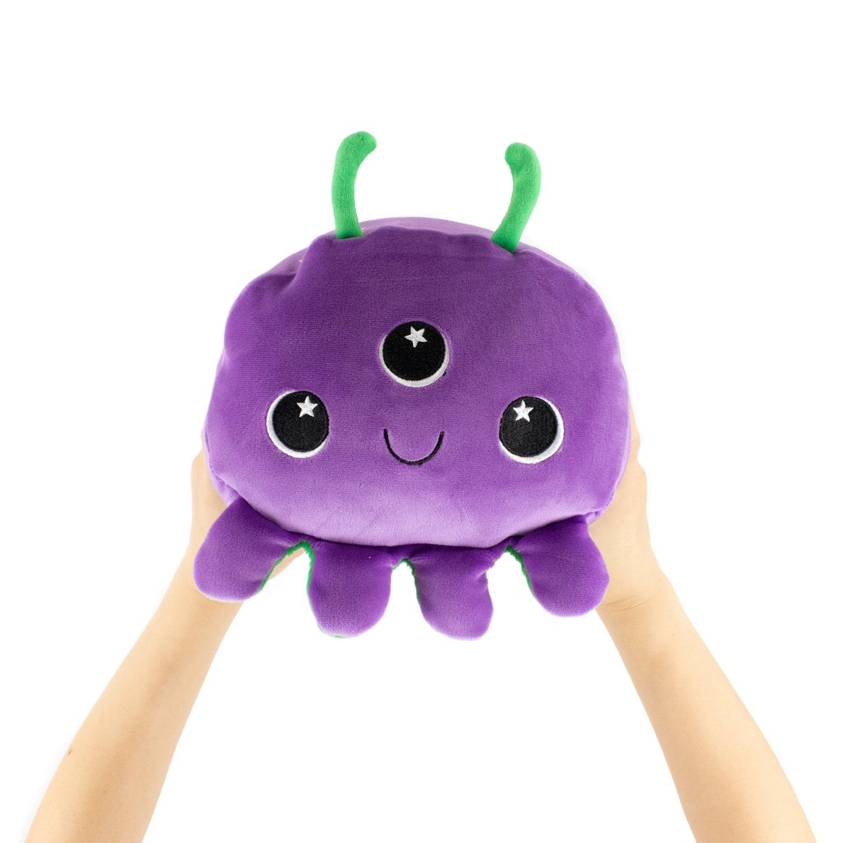 Two hands hold up Demian the Demon, a round purple plush toy with three starry eyes, a cute smile, green antennae, and short legs—an adorable alien-inspired stuffed animal perfect for your next cuddle buddy.