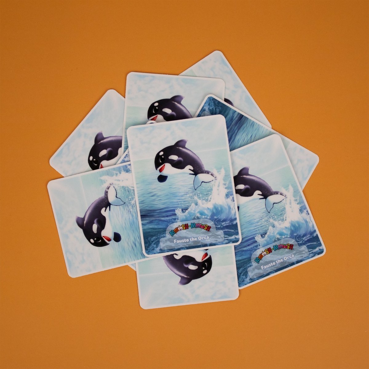 Seven Moosh-Moosh 3" x 4" vinyl stickers show an orca leaping from the water, scattered on an orange surface. Each sticker features the text "Fauste the Orca.