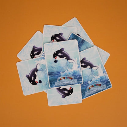 Seven Moosh-Moosh 3" x 4" vinyl stickers show an orca leaping from the water, scattered on an orange surface. Each sticker features the text "Fauste the Orca.
